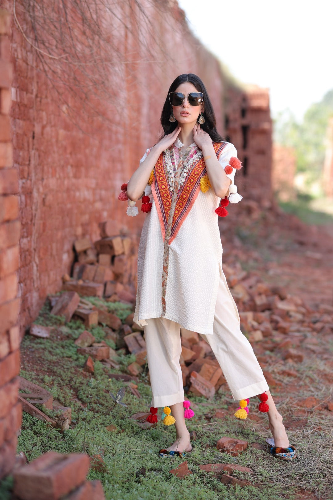 Model wearing ivory kurta set with tribal embroidery and tassels, standing against a red brick backdrop.
