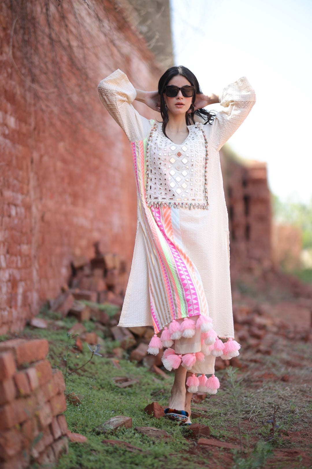 Model in ivory kurta with mirror detailing and vibrant tassels, standing against rustic brick wall.