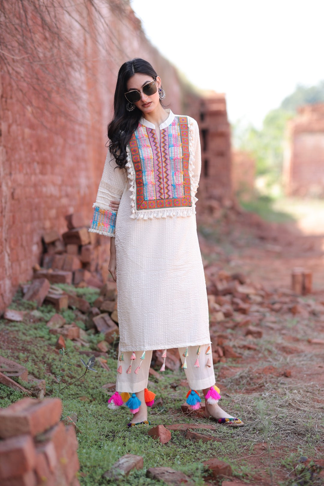 Model wearing ivory kurta with colorful embroidered patch and tassel pants in rustic outdoor setting