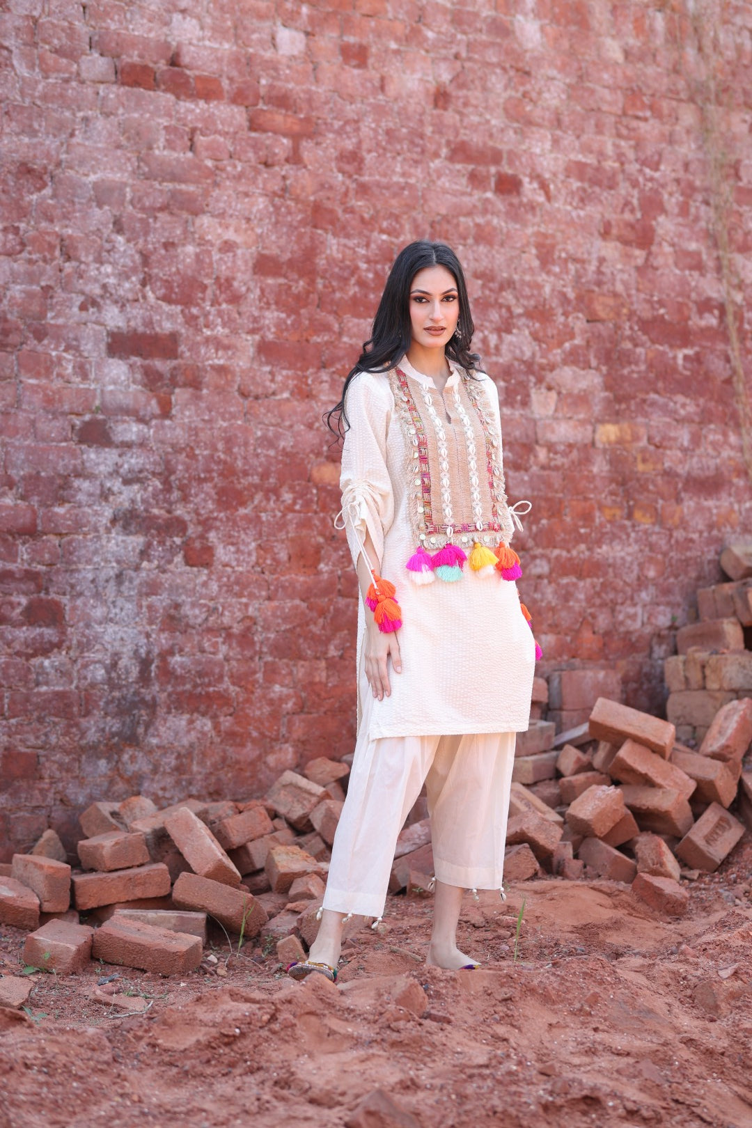 Model wearing ivory textured kurta with multicolour tassels and mirror detailing, standing against a rustic brick backdrop.