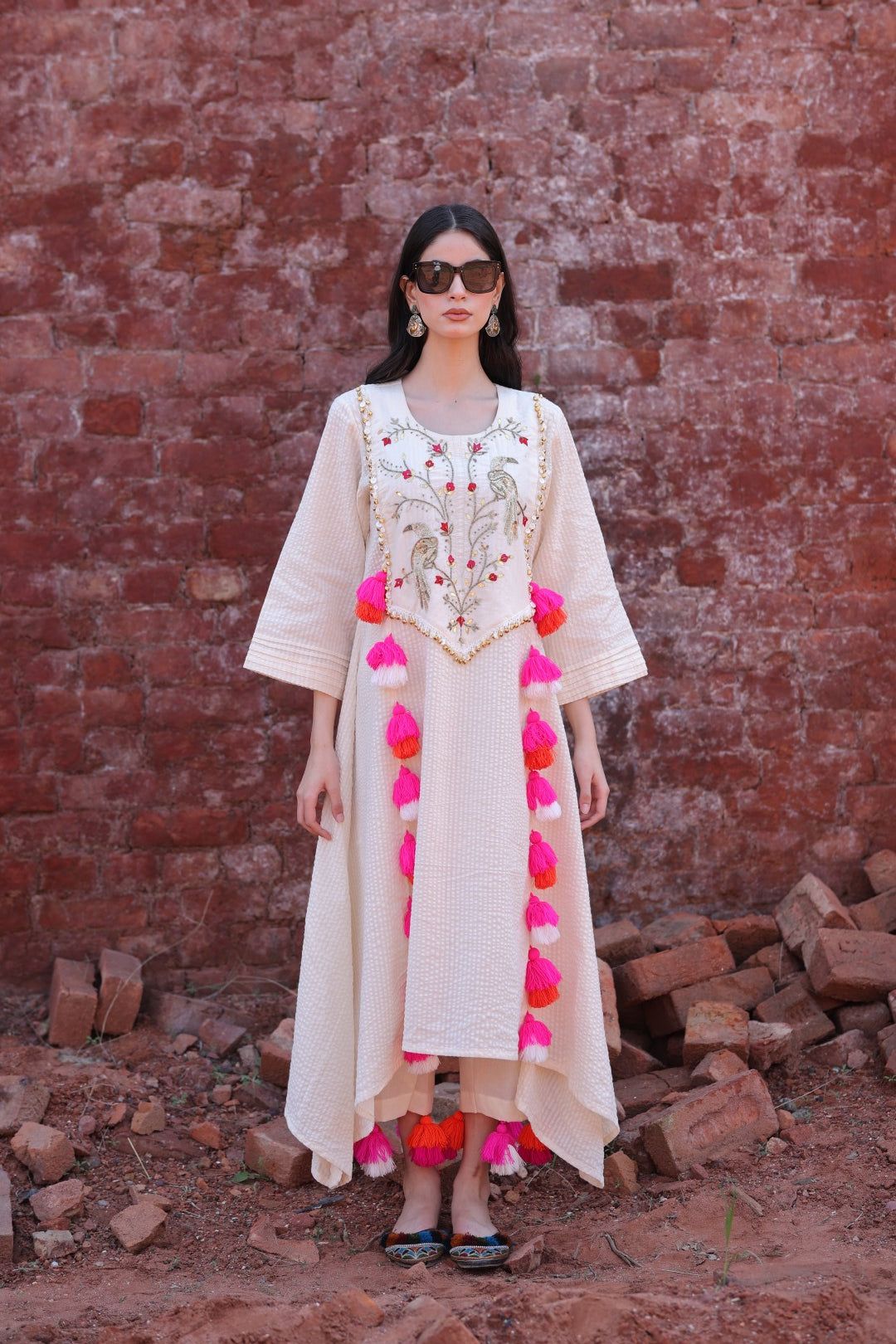 Model in ivory kurta with parrot embroidery and multicolour tassels, standing against a rustic brick wall