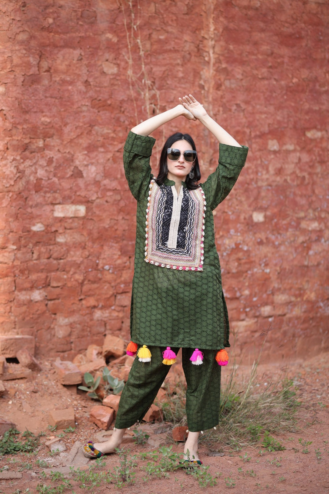 Model wearing deep green embroidered kurta set with colourful tassels and shell yoke, standing against rustic wall
